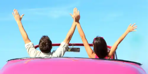 A picture of a couple in a pink car, with their hands up in excitement.